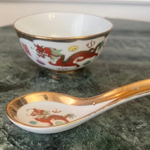 Könnte beinhalten: Eine weiße Porzellanschale und ein Löffel mit goldenen Verzierungen und einem roten Drachenmuster. Schale und Löffel befinden sich auf einer grünen Marmorfläche.