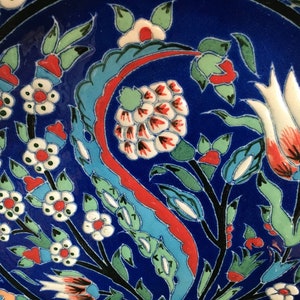 May include: A blue ceramic plate with a floral design. The design features white, red, and green flowers and leaves. The plate has a black border.