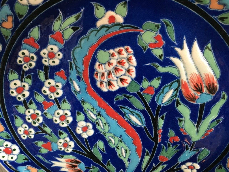 May include: A blue ceramic plate with a floral design. The design features white, red, and green flowers and leaves. The plate has a black border.