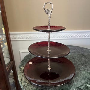 Puede incluir: Un soporte de servicio de tres niveles con platos de vidrio de color rojo oscuro de diferentes tamaños. La columna central de soporte es de metal plateado con un diseño texturizado similar a una rama y un asa decorativa en forma de hoja.