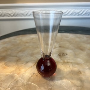 Può includere: Un bicchierino da shot in vetro trasparente con una base rotonda di colore rosso scuro. Il bicchiere è alto e stretto, con un bordo svasato.