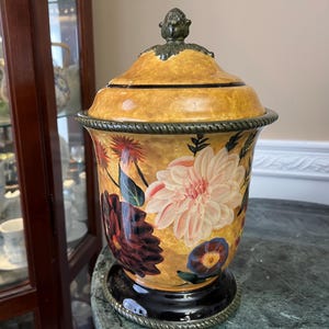 Puede incluir: Un tarro decorativo de cerámica con tapa, con un diseño floral en tonos rosa, rojo y azul sobre un fondo amarillo. El tarro tiene una base negra y un adorno verde en la tapa. El tarro mide aproximadamente 30 cm de altura.