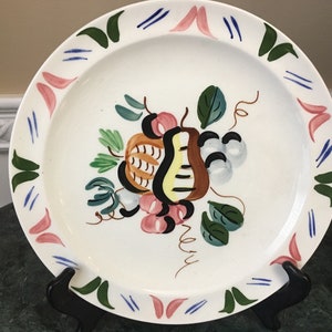 Könnte beinhalten: Ein weißer Keramik-Dinnerteller mit handbemaltem Design aus Früchten und Blättern in Grün-, Rosa-, Gelb-, Braun- und Schwarztönen. Der Teller hat einen dekorativen Rand mit rosa, grünen und blauen Akzenten.