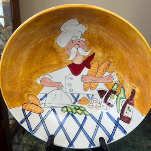 Könnte beinhalten: Eine große, runde Servierplatte aus Keramik mit handgemaltem Design. Das Bild zeigt einen Koch, der Baguettes hält, mit Weinflaschen, Käse und Trauben auf einem Tisch mit einer blau-weiß karierten Tischdecke. Der Hintergrund ist gelb.