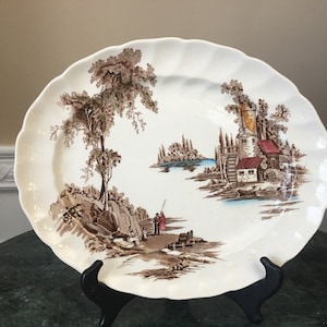 May include: A white ceramic plate with a scalloped edge, featuring a brown and white landscape scene. The scene depicts trees, buildings, and figures. The plate is displayed on a black stand.