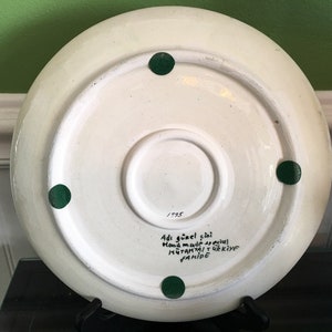 May include: White ceramic plate with green painted dots on the bottom. The plate has handwritten text in black ink that reads "Adi g&uuml;zel &ccedil;ini hand made special  MUTFAKTAN  TURKIYE  YAPIMI 1995"