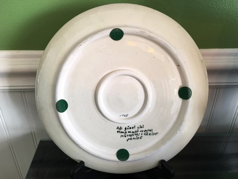 May include: White ceramic plate with green painted dots on the bottom. The plate has handwritten text in black ink that reads "Adi g&uuml;zel &ccedil;ini hand made special  MUTFAKTAN  TURKIYE  YAPIMI 1995"