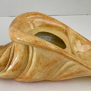 May include: A ceramic cornucopia planter in a light beige color with a speckled finish. The planter has a wide, open mouth and a long, curled horn.