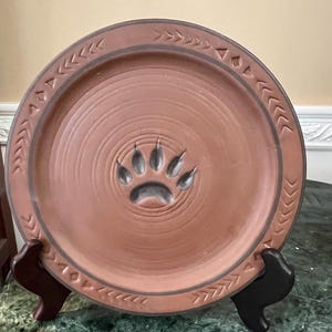 Puede incluir: Un plato redondo de cerámica color terracota con un diseño de huella de pata en el centro. El plato tiene un borde elevado con patrones de flechas y triángulos grabados. El plato se exhibe sobre un soporte negro.