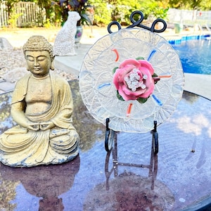 May include: A glass plate featuring a pink flower design is displayed on a black metal stand. The plate is clear with a textured surface and has a pink flower with white accents in the centre. The plate is surrounded by a white border with blue and orange accents. A golden Buddha statue is sitting on a table in the background.