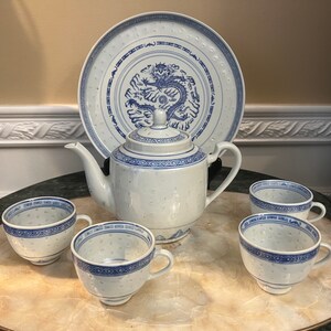 May include: A white porcelain tea set with blue accents. The set includes a teapot, four teacups, and a large plate. The plate features a blue dragon design. The teacups and teapot have a delicate dotted pattern.