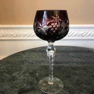 May include: A tall, clear wine glass with a deep red, cut-glass bowl. The bowl is decorated with a grapevine and grapes pattern. The stem is faceted and the base is clear.