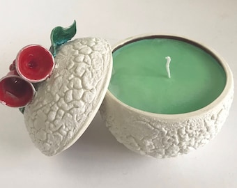 May include: White ceramic candle holder with a lid decorated with red holly berries and green leaves. The candle holder is filled with a green scented candle.