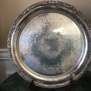 Op de afbeelding: Een grote, ronde, zilveren dienblad met een sierlijk, gegraveerd ontwerp. Het dienblad heeft een verhoogde rand en een glad, reflecterend oppervlak.