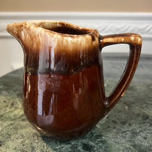 May include: A small, brown ceramic pitcher with a handle. The pitcher has a glossy finish with a gradient of colors, from a light tan at the top to a dark brown at the base. The handle is a solid brown color.