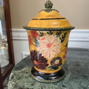 Puede incluir: Un tarro decorativo de cerámica con tapa. El tarro tiene una base y tapa amarillas, decorado con diseños florales en rojo, rosa y marrón. Tiene una base negra y ribete verde, lo que le da un aspecto vintage.
