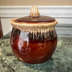 Puede incluir: Un tarro de cerámica marrón con tapa. El tarro tiene un acabado brillante con una base marrón oscuro y un diseño marrón claro goteado alrededor del borde. La tapa tiene un pomo en la parte superior.