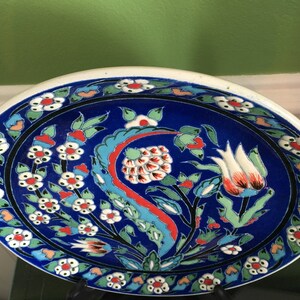 May include: A blue and white ceramic plate with a floral design. The plate has a white rim and a blue center with white flowers and red and green leaves. The plate is oval shaped and has a glossy finish.