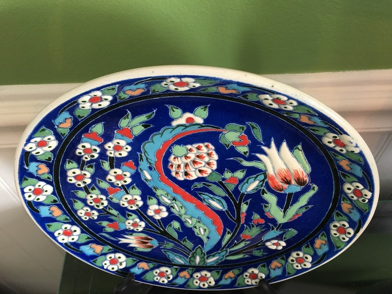 May include: A blue and white ceramic plate with a floral design. The plate has a white rim and a blue center with white flowers and red and green leaves. The plate is oval shaped and has a glossy finish.