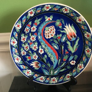 May include: A blue and white ceramic plate with a floral design. The plate features a central motif of a stylized tulip with red, white, and green accents. The border of the plate is decorated with a repeating pattern of white flowers with red centers.