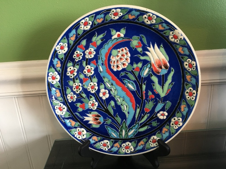 May include: A blue and white ceramic plate with a floral design. The plate features a central motif of a stylized tulip with red, white, and green accents. The border of the plate is decorated with a repeating pattern of white flowers with red centers.