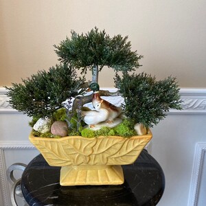May include: A yellow ceramic planter with a leaf design, featuring three small artificial trees and a ceramic figurine of two ducks. The planter is sitting on a black surface.