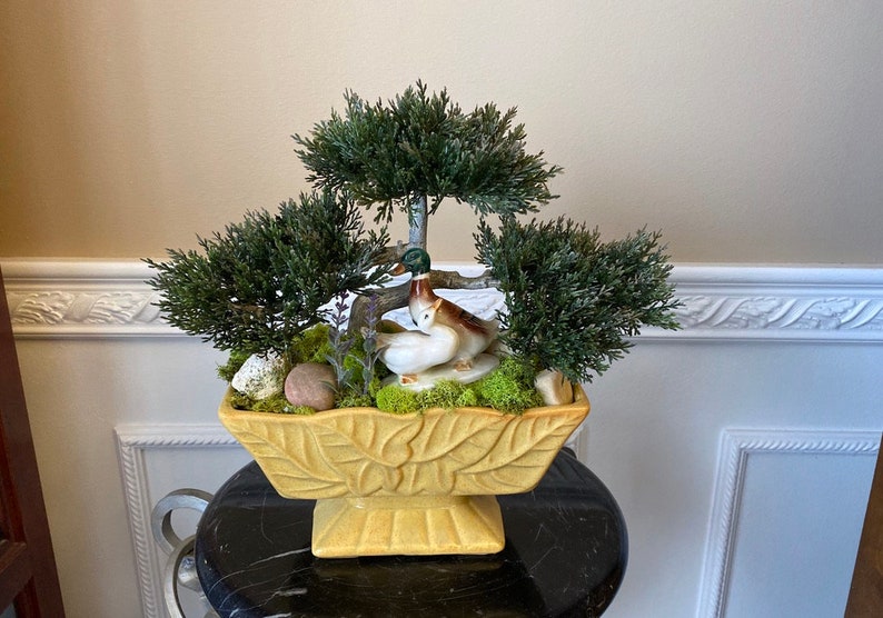 May include: A yellow ceramic planter with a leaf design, featuring three small artificial trees and a ceramic figurine of two ducks. The planter is sitting on a black surface.