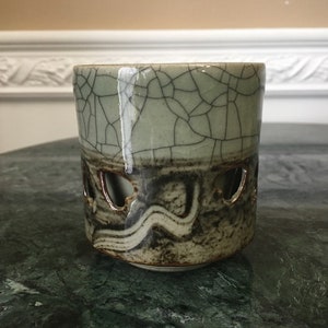 Op de afbeelding: Een keramische kandelaar met een lichtgroen, gecraqueleerd glazuur. Het onderste gedeelte heeft een donkerbruin en grijs ontwerp met hartvormige uitsparingen. De kandelaar is cilindrisch en staat op een groen oppervlak.