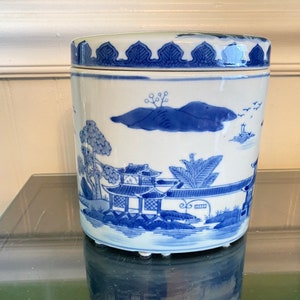 May include: A cylindrical, white ceramic jar with a lid, adorned with blue landscape illustrations. The jar features a detailed design of pagodas, trees, and mountains. The lid has a decorative blue border.
