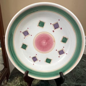 May include: A large, round ceramic serving bowl with a white interior. The bowl has a green and pink painted rim, with a central pink spiral design. The interior is decorated with green and purple square shapes and small red dots.