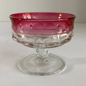 May include: A clear glass dessert dish with a pink rim and a textured pattern. The dish is on a clear glass pedestal.