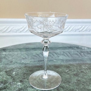 May include: A clear glass champagne coupe with a delicate floral etched design. The coupe has a long, slender stem and a wide, shallow bowl.