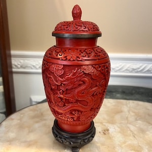 Peut inclure: Un vase chinois en laque de cinabre rouge avec un motif de dragon. Le vase a un couvercle et repose sur un piédestal en bois.