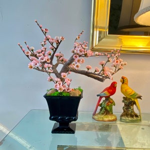Op de afbeelding: Een zwarte keramische pot met een bonsaiboom met roze bloesems. Twee keramische vogels, een rood en een geel, staan op een glazen tafel voor de pot.