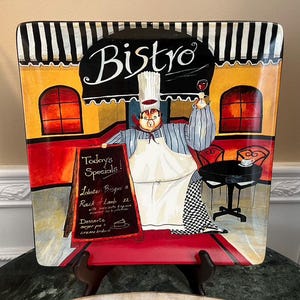 Könnte beinhalten: Eine farbenfrohe Keramik-Servierplatte mit einem Cartoon-Koch, der ein Glas Wein hält und eine Speisekarte mit der Aufschrift "Heute's Specials!" Die Platte zeigt eine Bistro-Szene mit einem rot-weiß-schwarz gestreiften Vordach und einem gelben Gebäude mit roten Fenstern.