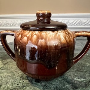 Puede incluir: Un azucarero vintage de cerámica marrón con tapa y dos asas. El cuenco tiene un acabado brillante con un efecto de esmalte goteado, mostrando tonos marrones. La tapa presenta un pequeño pomo para facilitar su manejo.