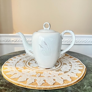 May include: White porcelain teapot with a lid and a curved spout. The teapot has a silver trim and a decorative design on the front. It sits on a round, yellow and white beaded placemat. The background is a neutral color.