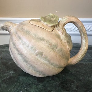 May include: A ceramic teapot shaped like a gourd with a green leaf lid. The teapot is glazed in shades of brown, green, and pink.