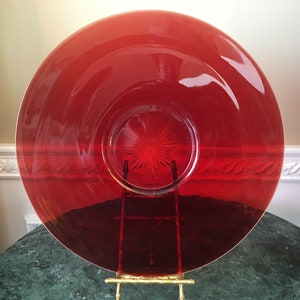 Könnte beinhalten: Ein großer, runder, roter Glasservierteller mit einem Sternmuster in der Mitte. Der Teller steht auf einem goldenen Metallständer.