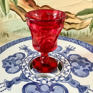 Puede incluir: Un pequeño gobelet de vidrio rojo con un diseño acanalado. El gobelet tiene un tallo y una base redonda. Está sentado sobre un plato de porcelana azul y blanco con un diseño decorativo.