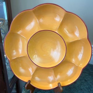 May include: A yellow ceramic serving platter with a flower-like design. The platter has a central bowl and several petal-shaped compartments. The rim is outlined in red. The platter is supported by a wooden stand.
