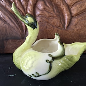 May include: A ceramic swan planter with a green and white glaze. The swan's neck is curved upwards and the planter has a wide, open mouth.