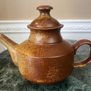Puede incluir: Una tetera de cerámica marrón con tapa y asa. La tetera tiene una textura moteada y un cuerpo redondeado. El pico está en ángulo y el asa es curva. La tetera probablemente se usa para preparar té.