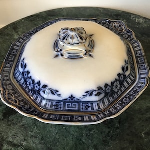 Könnte beinhalten: Eine weiß-blaue Keramik-Butterdose mit Deckel. Die Schale hat ein Blumen- und geometrisches Muster mit goldenem Rand. Der Deckel hat einen dekorativen Griff. Die Schale ist achteckig.