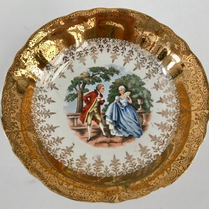 May include: A white porcelain saucer with a gold rim and a floral pattern. The center of the saucer features a painted scene of a couple in a garden setting.