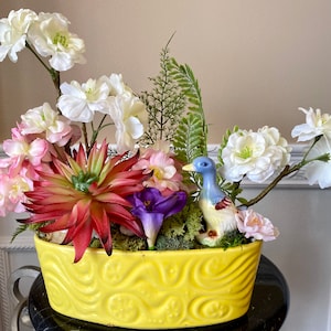 Könnte beinhalten: Ein gelber Keramiktopf mit einem wirbelnden Muster beherbergt eine Blumenarrangement. Die Anordnung umfasst rosa, weiße und rote Blumen, eine violette Blume, grünes Laub und eine blaue und weiße Keramik-Entenfigur.