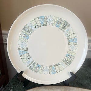 Op de afbeelding: Witte keramische plaat met een modern design uit het midden van de eeuw. De plaat heeft een ring van geometrische vormen in blauw, grijs en groen langs de rand. De plaat wordt op een zwarte standaard gepresenteerd.