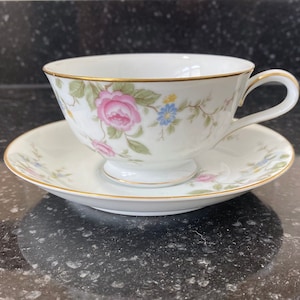May include: A white teacup and saucer set with a floral pattern featuring pink roses, blue flowers, and green leaves. The cup has a gold rim and a delicate handle.
