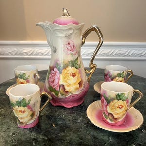 May include: A vintage porcelain tea set with a floral design. The set includes a teapot, creamer, sugar bowl, and four teacups and saucers. The floral design features pink and yellow roses.