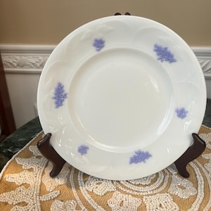 Puede incluir: Plato de cerámica blanca con un diseño floral en relieve y detalles en lavanda. El plato se exhibe sobre un soporte de madera marrón, resaltando su elegante diseño. Una pieza decorativa para servir o exhibir.